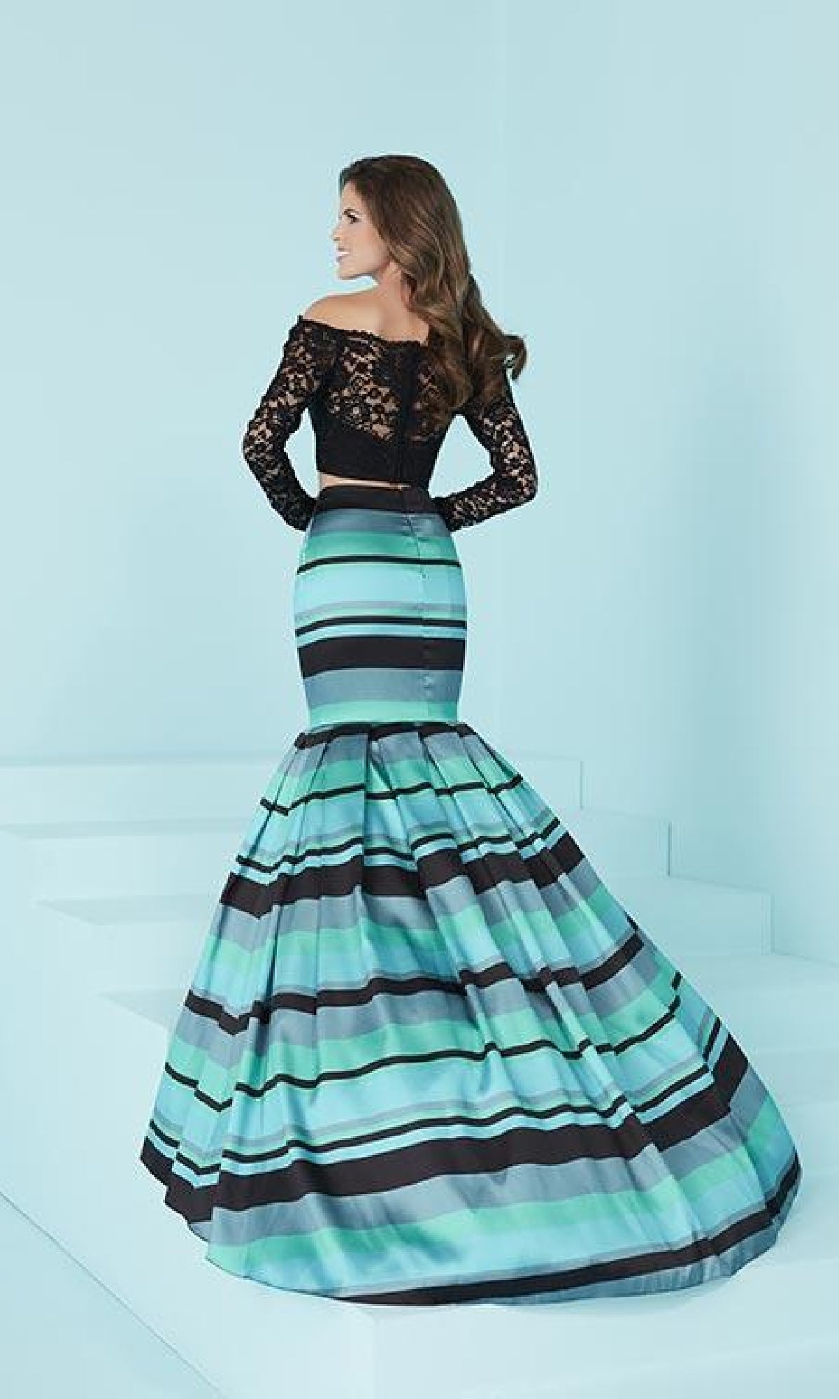 A woman stands on white steps wearing the Two-Piece Striped Mermaid Prom Dress, featuring a black lace long sleeve top and a bold mermaid skirt with blue and black horizontal stripes.
